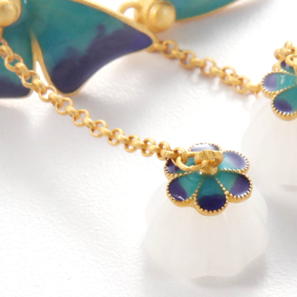 Close-up of Aveera designer butterfly wing drop earring showing intricate blue enamel work, gold chain, and white pendant.