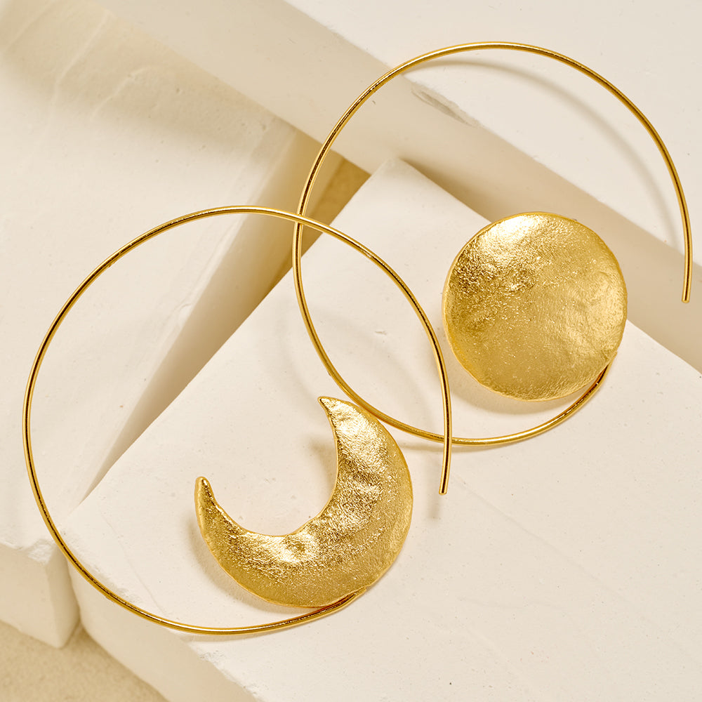 Pair of gold asymmetrical sun and moon large hoop earrings on white background.