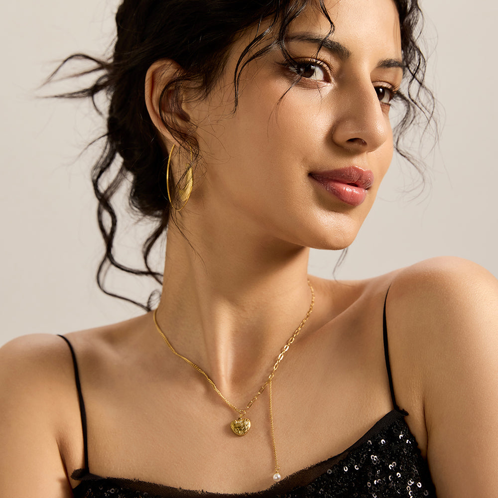 Model posing wearing gold tone textured heart and pearl dangle necklace.
