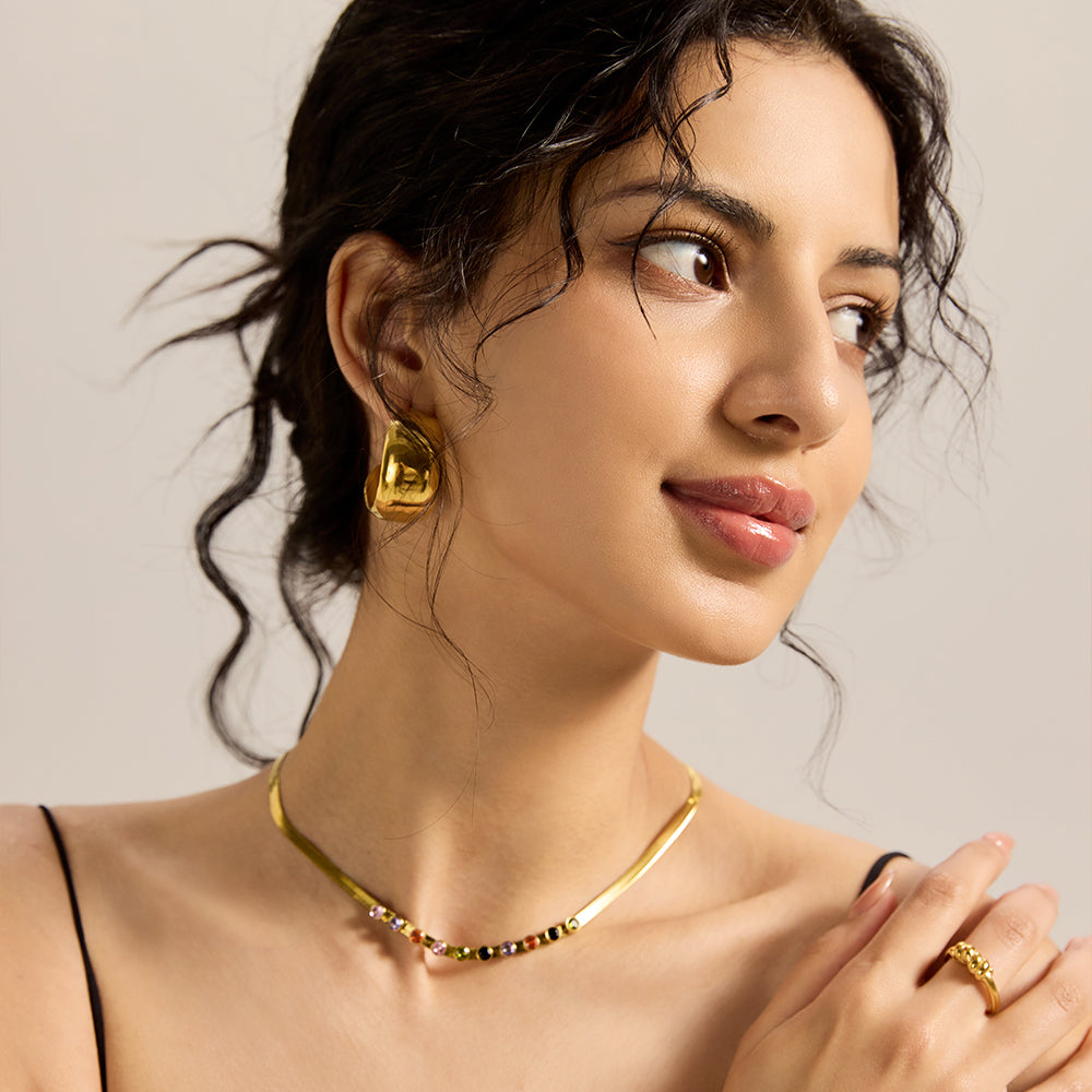 Model wearing gold snake chain necklace with multicolor gemstones, styled with matching earrings.
